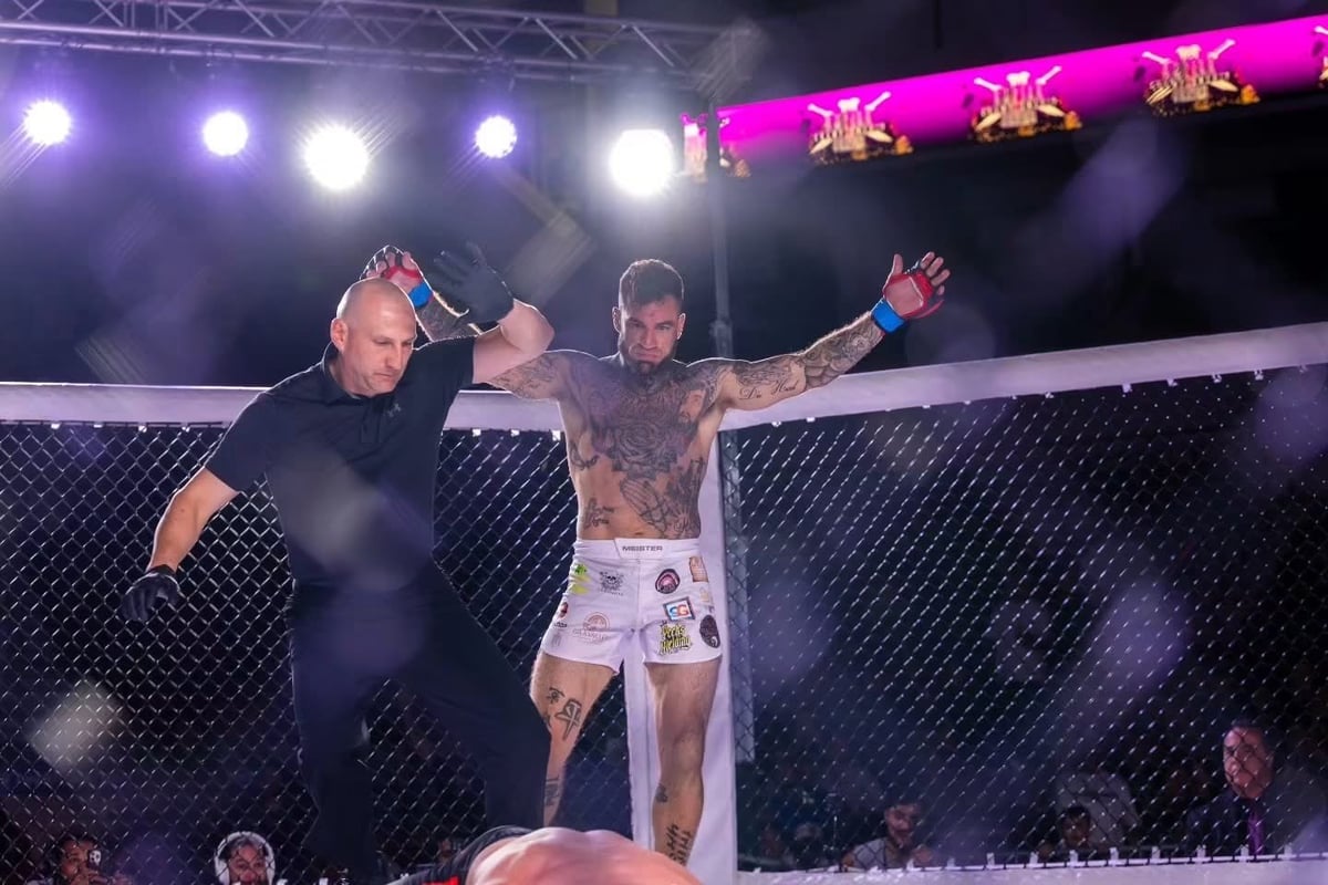 MMA fighter celebrating victory with arms raised inside octagon cage with referee and crowd in background