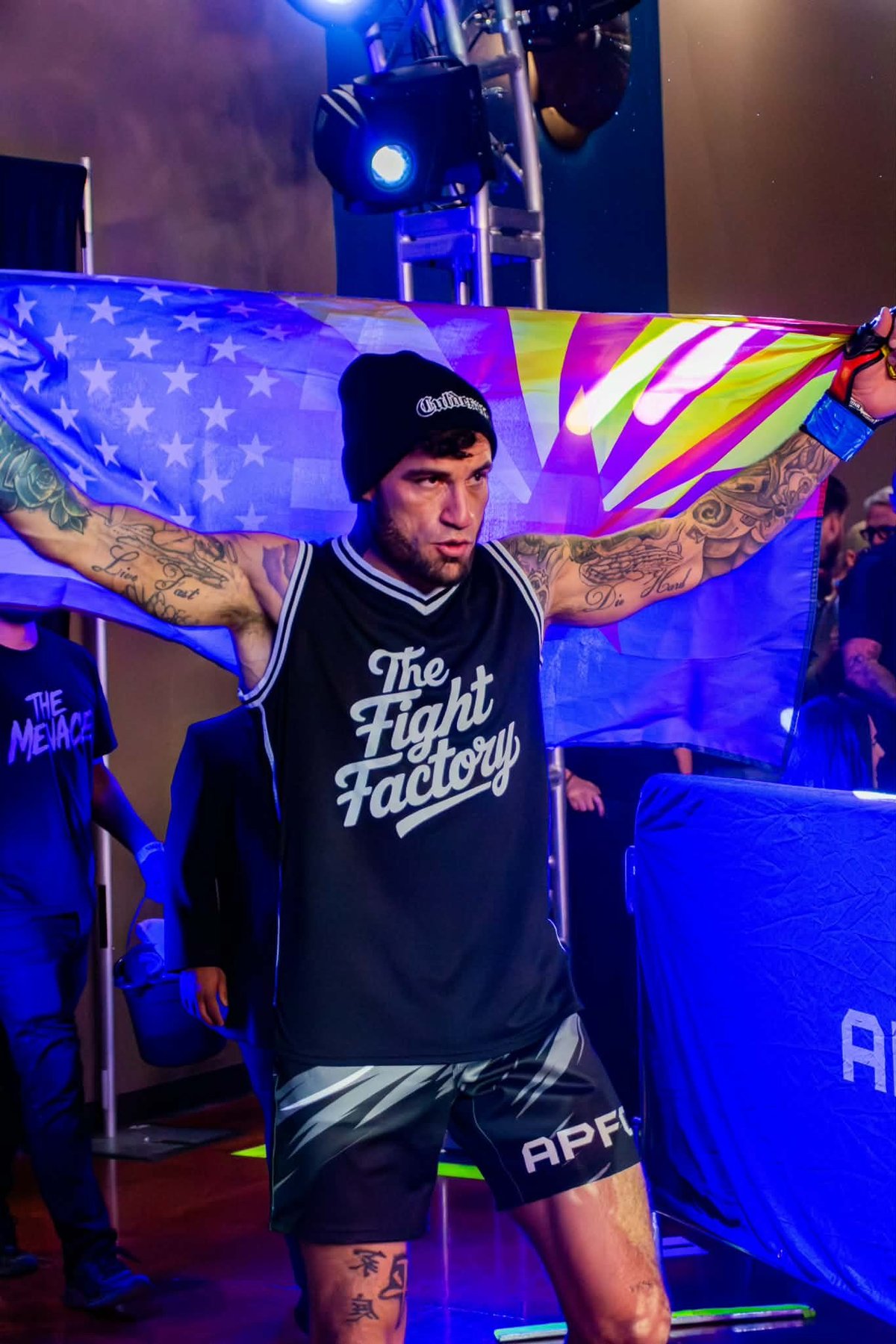 Tattooed performer on stage wearing black The Fight Factory vest and shorts, holding colorful American flag while blue stage lights illuminate the venue