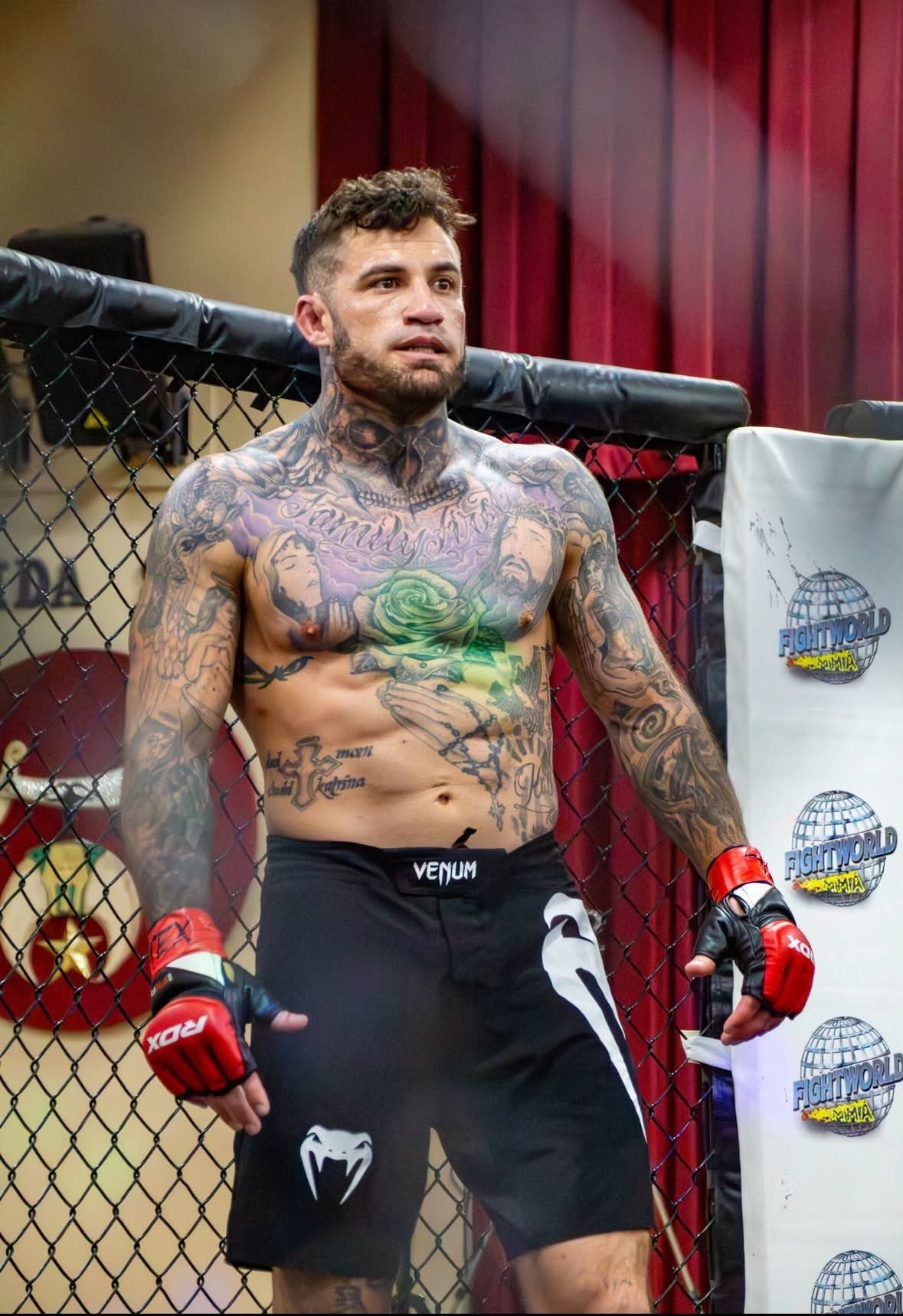 Tattooed MMA fighter leaning against octagon cage wearing red and white gloves and Venum shorts