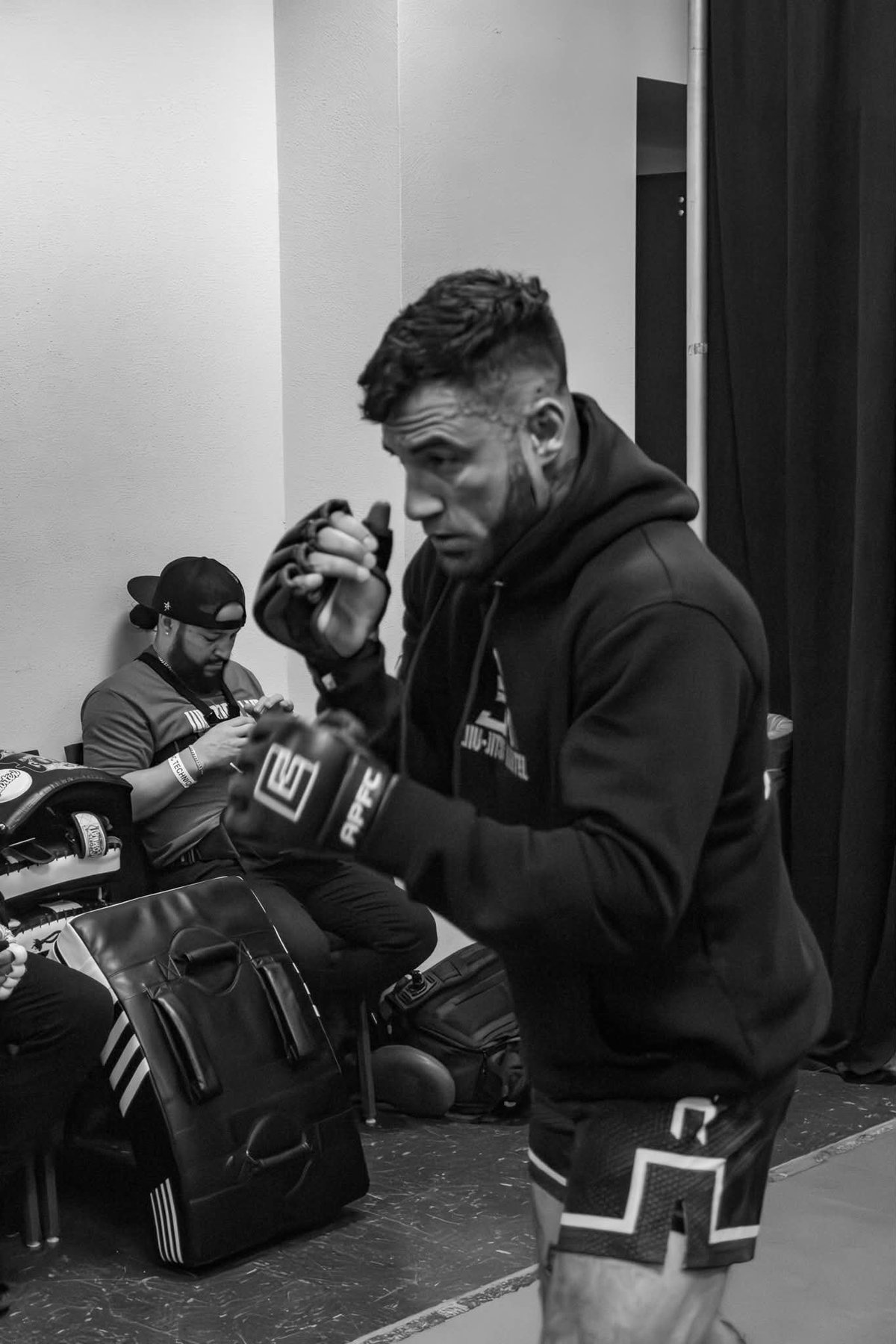 Black and white photo of a man in boxing gloves and hoodie with another person preparing equipment in the background