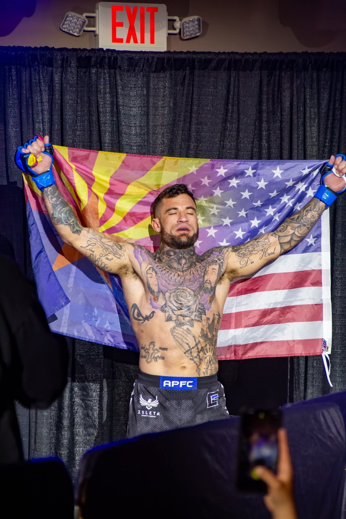 Tattooed fighter with arms raised holding Arizona and American flags on stage under red EXIT sign